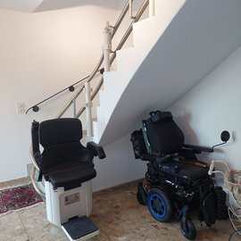 Sitz eines Treppenlifts, der eine gewinkelte Treppe hochführ, daneben ein Elektrorollstuhl
