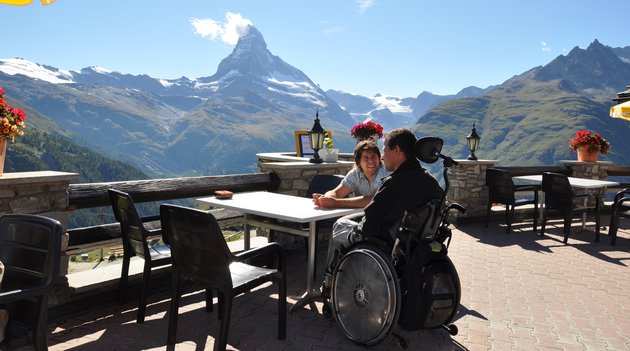 Zwei junge Männer, einer im Rollstuhl, sitzen auf einer Terrasse vor dem Matterhorn