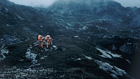 Foto von einer Berglandschaft. Zwei Männer, die wie Astronauten gekleidet sind, gehen zwischen Felsen umher.