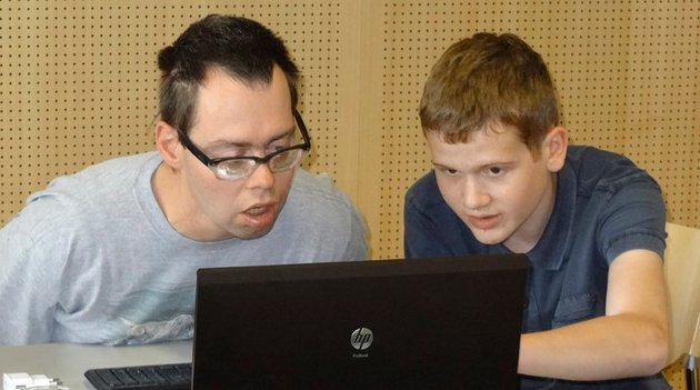 Zwei jungen Männern hinter einem Laptop, einer erklärt etwas. 