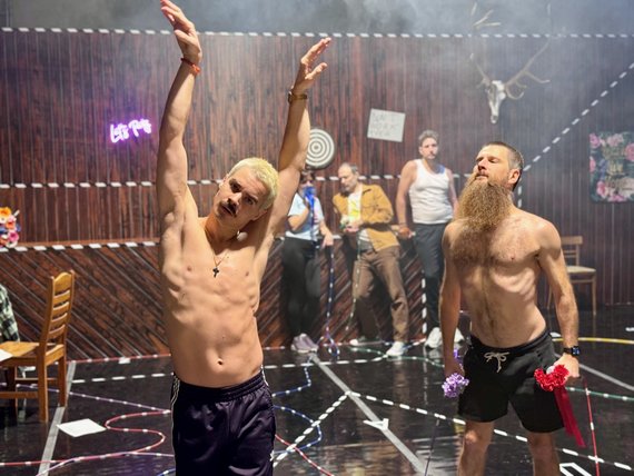 Zwei Tänzer in Shorts und mit nacktem Oberkörper. Im Hintergrund stehen drei Männer und schauen den Tänzern zu.
