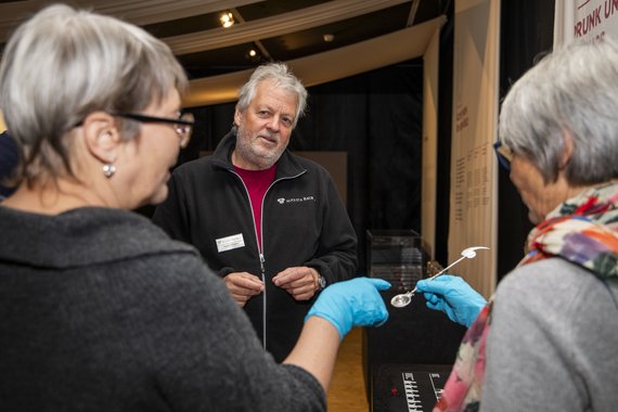 Zwei Frauen, von seitlich hinten fotografiert. Ihnen gegenüber steht ein Mann. Die beiden Frauen tragen Kunststoffhandschuhe. Eine Frau hält einen Gegenstand. Die andere Frau ertastet ihn mit dem Finger.