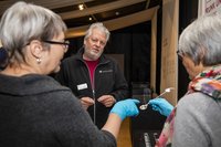 Zwei Frauen, von seitlich hinten fotografiert. Ihnen gegenüber steht ein Mann. Die beiden Frauen tragen Kunststoffhandschuhe. Eine Frau hält einen Gegenstand. Die andere Frau ertastet ihn mit dem Finger.