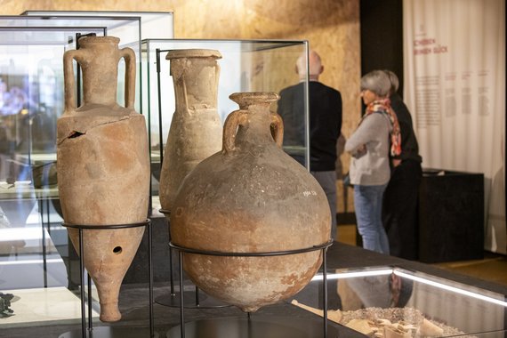 Foto: In einem Raum stehen im Vordergrund in Haltern drei Tongefässe mit unterschiedlichen Formen und Grössen. Im Hintergrund betrachten drei Besuchende Ausstellungsobjekte.