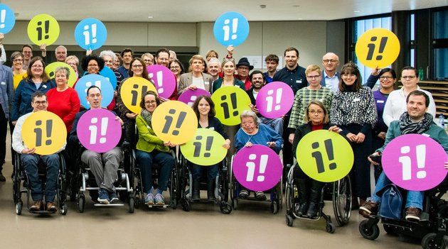 Menschen mit und ohne Behinderungen, die das Logo der Inklusionsinitiative in die Luft halten