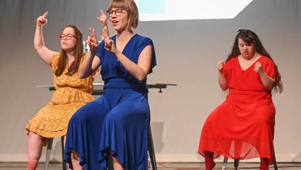 Drei Frauen in farbigen Röcken sitzen auf Stühlen und machen verschiedene Handzeichen