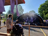 Foto von 2 Besuchern des Festivals. 1 Besucher sitzt im Rollstuhl. Sie schauen und hören sich ein Konzert an. 