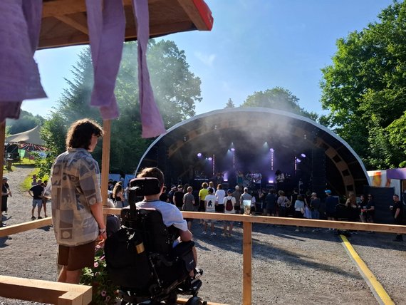 Foto von 2 Besuchern des Festivals. 1 Besucher sitzt im Rollstuhl. Sie schauen und hören sich ein Konzert an. 