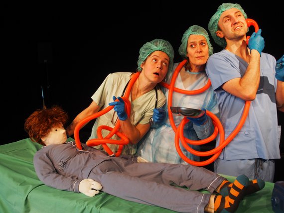 Zwei Schauspielerinnen und ein Schauspieler sind als OP-Team verkleidet. Ein langer Schlauch ist um ihre Hälse gelegt. Ein Ende des Schlauches hält der Mann an sein Ohr, das andere Ende führt in den Oberkörper einer Puppe, die vor ihnen auf dem OP-Tisch liegt.