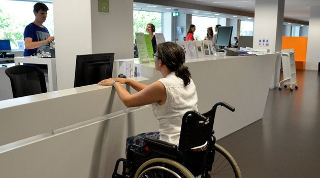 Eine Frau sitzt im Rollstuhl vor einem Schalter eines Büros.