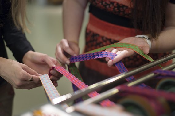 Foto einer Seidenbandmaschine. Zwei Frauen halten verschiedenfarbige Seidenbänder in den Händen. Die Bänder sind in unterschiedlichen Mustern gewoben.