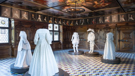 Foto: In einem historischen Zimmer mit Malereien an der Decke und den Wänden stehen fünf Puppen, die Kleider aus der Zeit von vor 400 Jahren tragen. Vier davon tragen weisse Frauenkleider. Eine Puppe ist ein Edelmann mit Schwert.