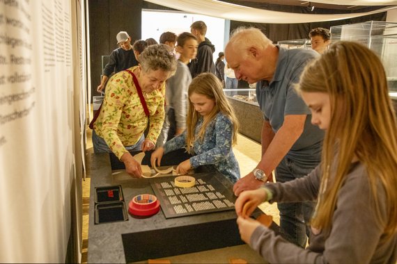 Menschen in einem Ausstellungsraum. Im Vordergrund stehen eine alte Frau, ein alter Mann und zwei Mädchen. Sie kleben Tonschalenteile zusammen.