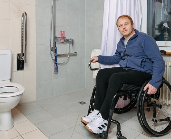 Ein Rollstuhlfahrer präsentiert sich stolz in seinem angepassten Badezimmer mit bodenebener Dusche