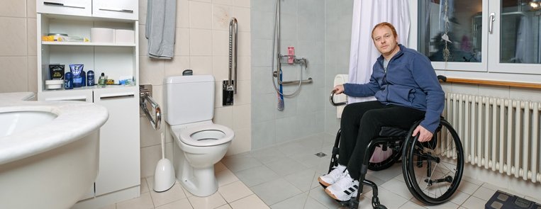 Ein Rollstuhlfahrer präsentiert sich stolz in seinem angepassten Badezimmer mit bodenebener Dusche