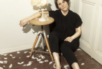 Photo copyright Margaux Corda : Une femme assise à même le sol, contre une porte. Elle est vêtue d'un tshirt et pantalon noir, ses genoux sont repliés. L'un de ses bras est posé sur un tabouret en bois sur lequel on devine une colombe empaillée. L'autre bras repose sur son genou. Son visage exprime une forme de lassitude. Le sol est jonché de quelques plumes.