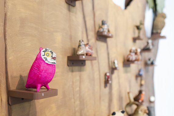 An einer Holzwand stehen auf kleinen Brettern verschiedene Skulpturen. Sie sind alle braun-beige. Auf einem Holzbrett steht eine Eule. Sie ist pink.