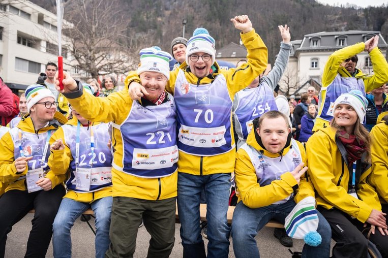  Des skieurs vêtus de combinaisons jaunes et de gilets bleus portant leur numéro de dossard applaudissent devant la caméra.